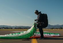 Pilote expérimenté vérifiant l'aile de son parapente près d'un aérodrome, avec montagnes au loin.