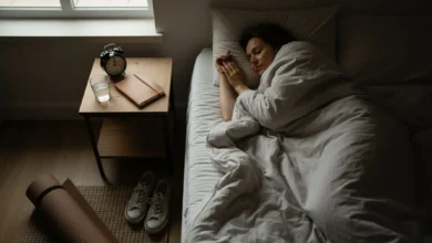 Photo en plongée d’une personne adulte endormie dans une chambre sombre mais chaleureuse, éclairée par une lumière douce venant de la fenêtre. Sur la table de nuit, un réveil analogique indique une heure de nuit avancée, à côté d’un verre d’eau et d’un carnet. La personne dort paisiblement, couette légèrement remontée, donnant une impression de bien-être et de repos profond. Un cadrage large laisse voir un tapis de yoga roulé et une paire de baskets au sol, suggérant un mode de vie sain lié à un bon sommeil.
