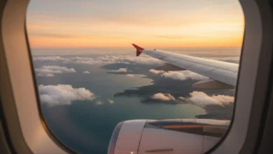 Vue aérienne spectaculaire prise depuis le hublot d’un avion, montrant une mer turquoise, une côte découpée et quelques nuages cotonneux. Au premier plan, on distingue le bout de l’aile de l’appareil. La lumière est dorée, évoquant un lever ou coucher de soleil, et l’ensemble donne une impression de liberté, de vacances et de voyage touristique.