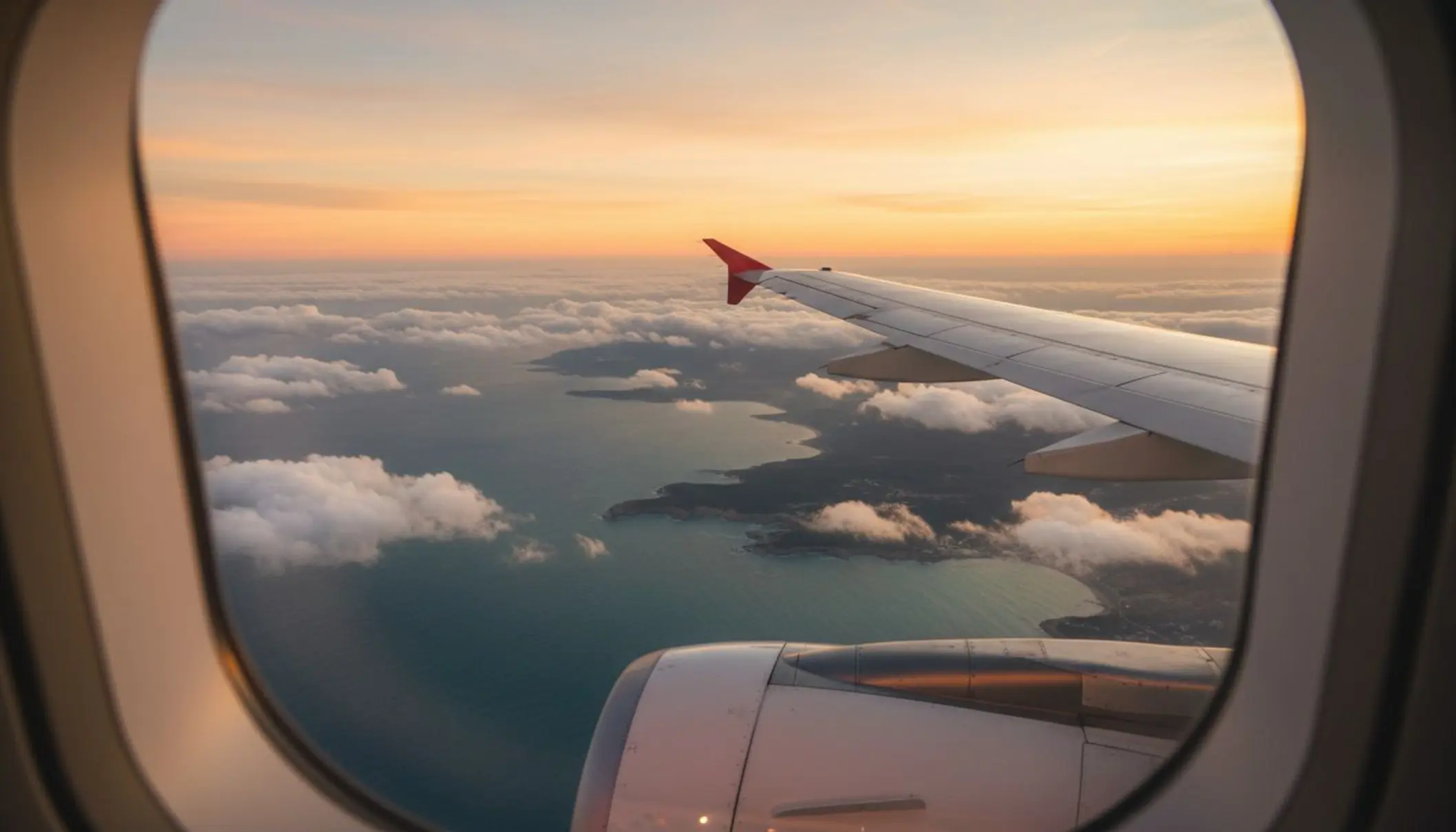 Vue aérienne spectaculaire prise depuis le hublot d’un avion, montrant une mer turquoise, une côte découpée et quelques nuages cotonneux. Au premier plan, on distingue le bout de l’aile de l’appareil. La lumière est dorée, évoquant un lever ou coucher de soleil, et l’ensemble donne une impression de liberté, de vacances et de voyage touristique.