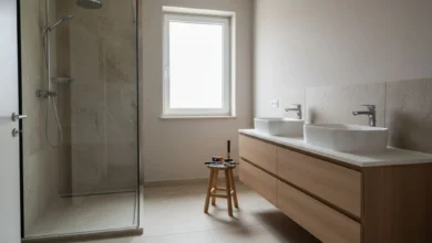 Photo lumineuse d’une salle de bains fraîchement rénovée, ambiance contemporaine chaleureuse : grande douche à l’italienne vitrée, meuble vasque en bois clair avec deux vasques blanches, carrelage imitation pierre au sol, mur peint dans une teinte douce, quelques outils de bricolage posés sur un tabouret en bois suggérant la fin du chantier. Lumière naturelle entrant par une fenêtre, atmosphère propre et apaisante qui illustre la réussite d’une rénovation.