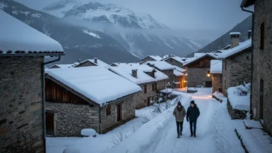 Photo en plongée douce d’un petit village de montagne français, toits enneigés, quelques lumières jaunes aux fenêtres au crépuscule. Au premier plan, deux personnes en doudoune marchent sur un chemin bordé de neige, sans foule, avec en arrière-plan un massif montagneux légèrement dans la brume. Ambiance chaleureuse, calme et intimiste, loin des grands domaines skiables.