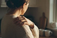 Main de femme pressée contre le haut du dos, geste de douleur dans un intérieur domestique éclairé naturellement