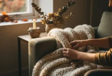 Mains disposant un plaid en grosse maille sur un canapé beige, lumière automnale dorée