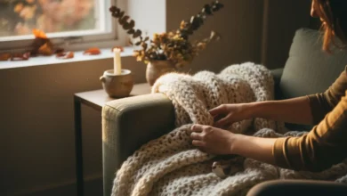 Mains disposant un plaid en grosse maille sur un canapé beige, lumière automnale dorée